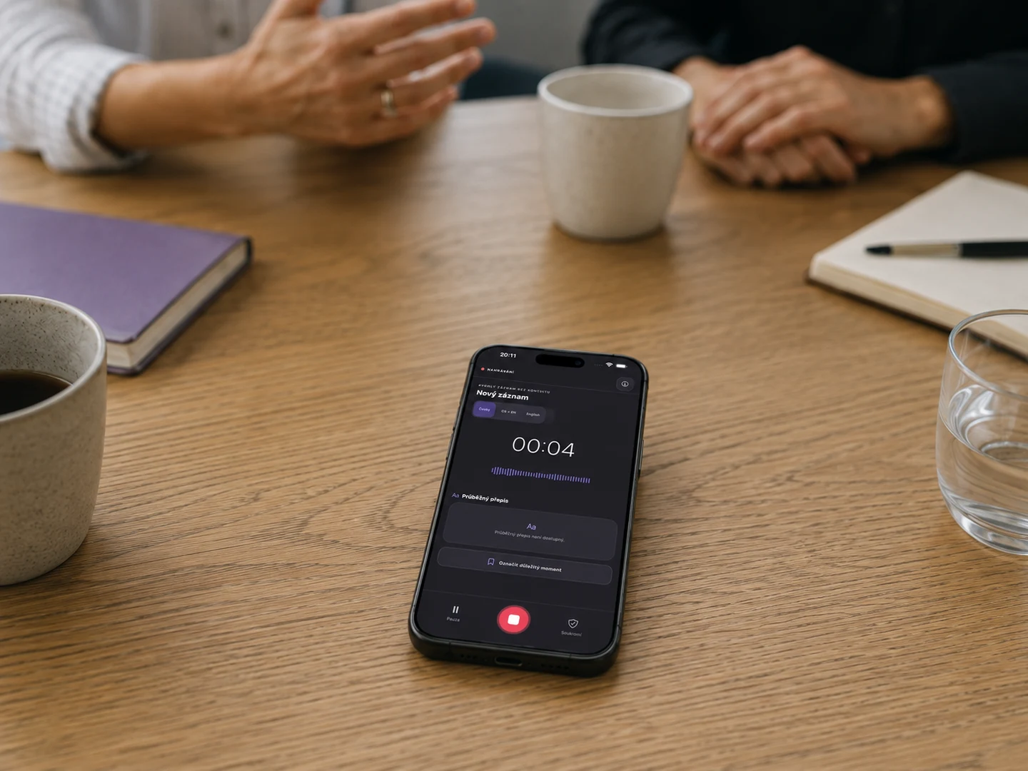 Telefon leží na stole a nahrává v Catch Minutes. Pohled šikmo shora ukazuje displej; kolem stolu probíhá rozhovor.